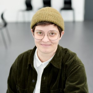 Es ist ein Potrait einer weißen Person. Der Oberkörper und der Kopf sind zu sehen sowie ein heller fast leerer Raum im Hintergrund mit grauem Boden und ein paar schwarzen Stühlen. Die Person trägt eine große Brille und eine hell-oliv-farbende Strickmütze. Unter der kurzen Mütze sind kurze dunkle Haare und beide Ohren zu sehen. Die person lächelt ganz leicht mir geschlossenem Mund. Die braunen Augen schauen direkt in die Kamera. Die person trägt ein weißes zugeknöptes Hemd und darüber eine offene dukelgrüne Cordjacke.