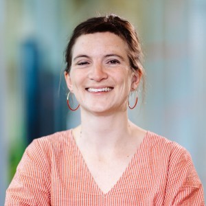 A mid-aged woman with dark hair and a red and white striped dress and round earrings has her arms crossed and smiles into the camera.